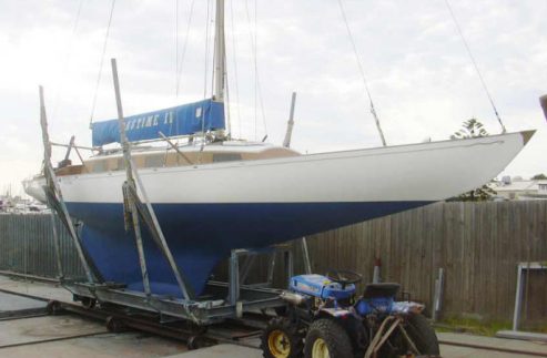 A blue and white classic sailing yacht rests on a cradle near a wooden fence, with a blue tractor parked in front. The sky is overcast.