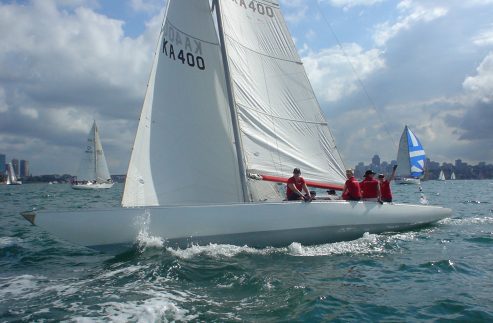 A white Square Metre Yacht with a crew of four in red shirts sails on choppy blue water. Other Skerry Cruisers are visible in the background beneath a partly cloudy sky.