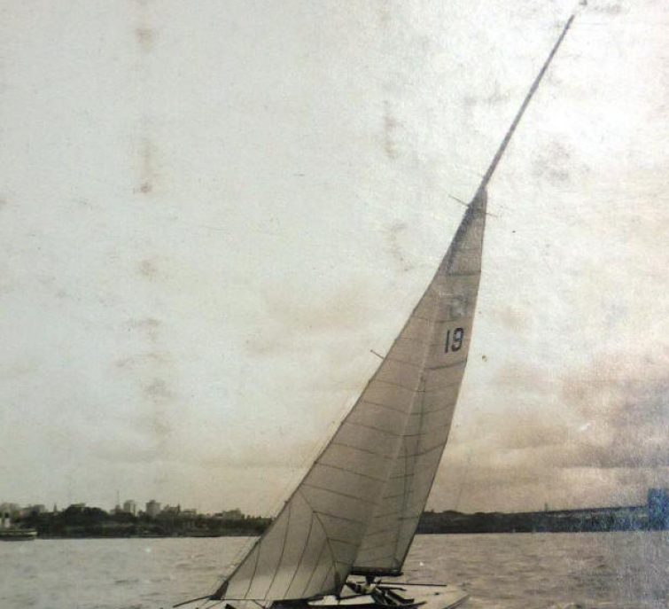 A black and white photo of a classic sailing yacht with the number 19 on its sail tilting to one side on the water. Bacchante is handwritten across the top. Trees and buildings are visible on the distant shore.