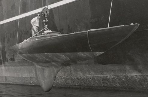 A man stands on a sleek Schärenkreuzer racing sailing boat being hoisted by a crane beside a large ship, with the classic sailing yacht partially above the water and ropes securing it from above.