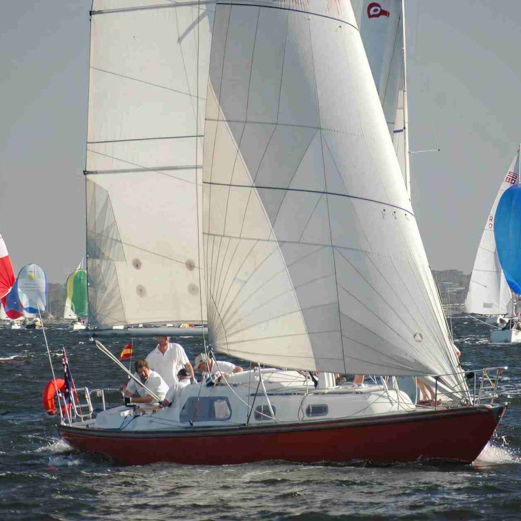 A red-and-white Classic Sailing Yacht with several people aboard sails on choppy water, surrounded by other colourful sailboats with bright spinnaker sails, under a clear sky.