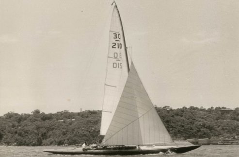 A vintage Skerry Cruiser with three people on board glides across the water, its sails marked 30, 210, and 015. Trees and a few buildings line the shore in the background beneath a clear sky.
