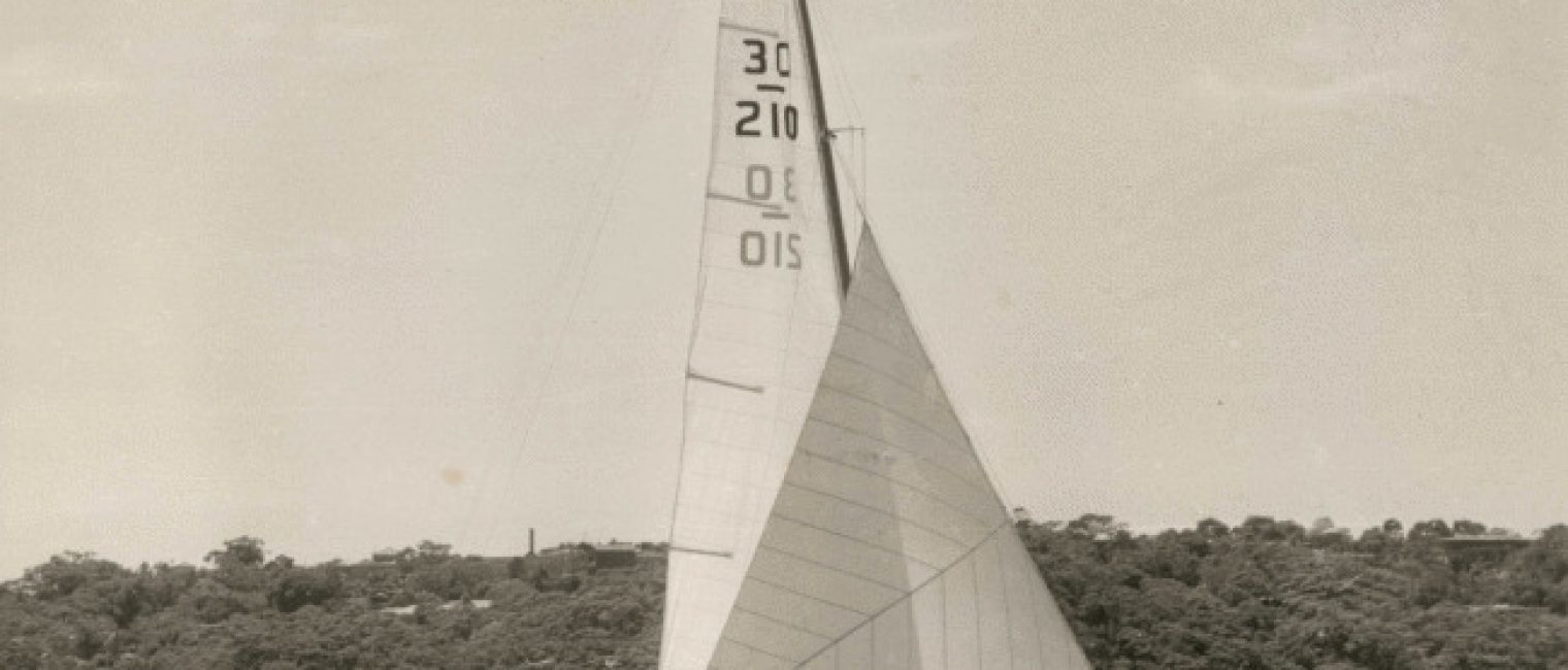 A vintage Skerry Cruiser with three people on board glides across the water, its sails marked 30, 210, and 015. Trees and a few buildings line the shore in the background beneath a clear sky.