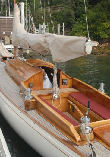 A wooden Skerry Cruiser with a covered mast is moored beside a pier, with a boathouse and trees in the background. The boat features polished wood details and red bench seating in the cockpit.