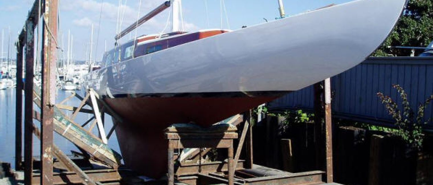 A white Square Metre Yacht with a wooden deck sits on a metal cradle on rails near a dockyard, ready to be launched into the water. The blue sky is dotted with clouds, and other boats are visible in the background.