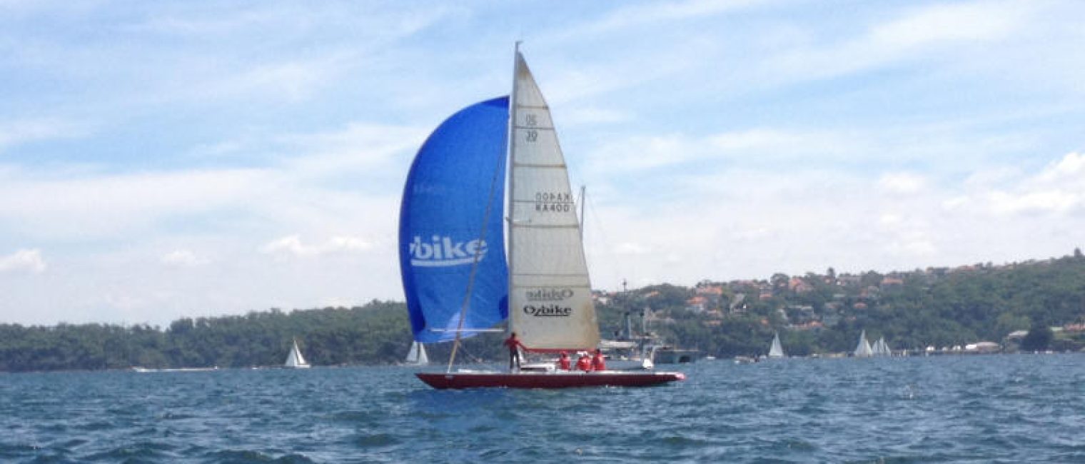 A classic sailing yacht with a large blue spinnaker and white sails glides across the water, with several people on board. Other Square Metre Yachts and a tree-covered shoreline with buildings are visible in the background.